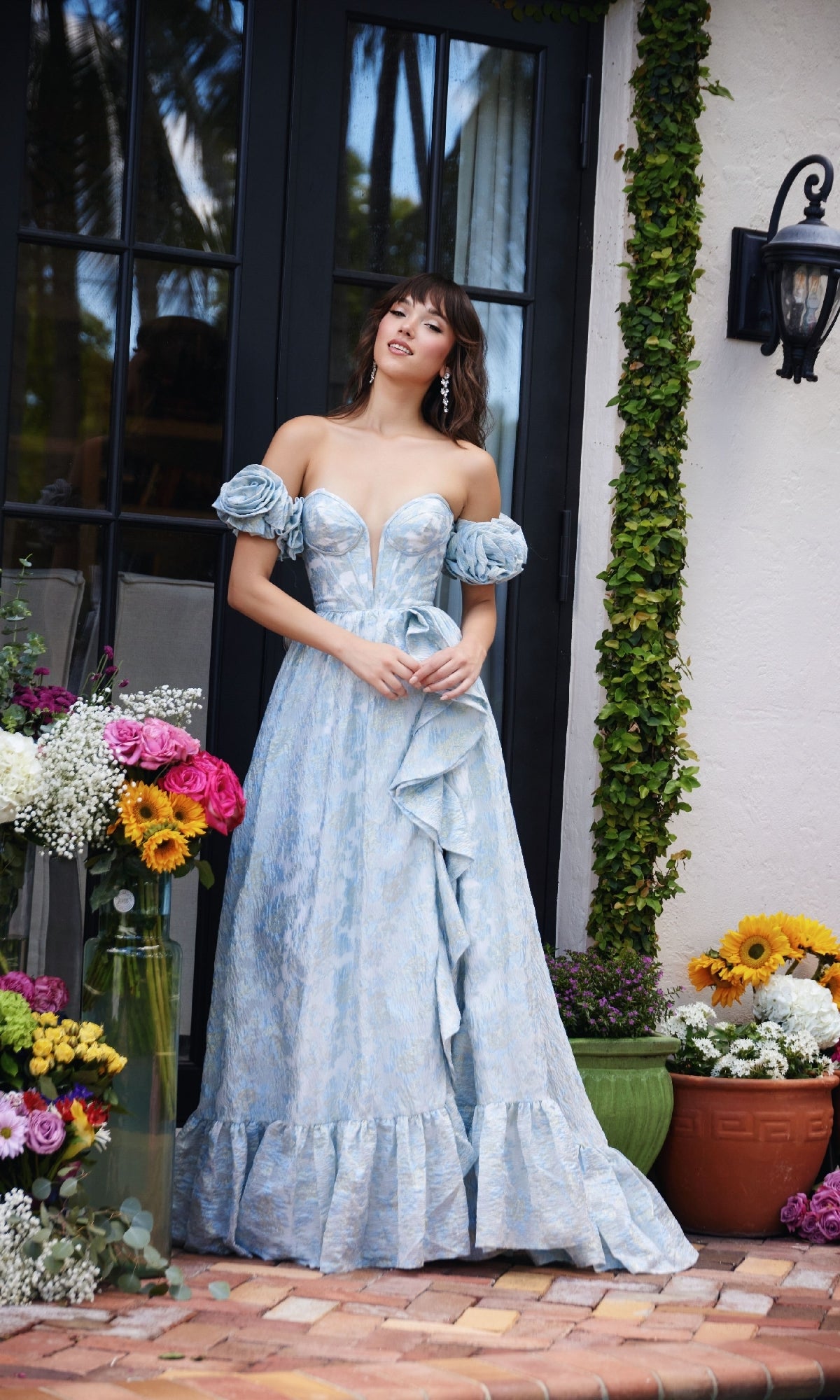A woman wearing the Ava Presley 40035 long formal dress, featuring a light blue gown with detached puff sleeves and a corset bodice, stands before a glass door amid lush flower arrangements.